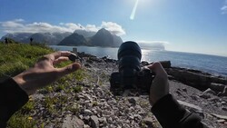 Photographer's hands holding a camera on a rocky beach with snow-capped mountains across the water.
