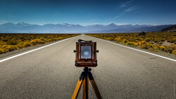 Large format camera on tripod centered on a straight desert highway with mountains in the distance.
