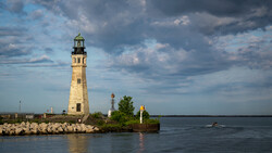 Stone lighthouse with green cupola on rocky breakwater under dramatic cloudy sky.