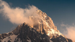 Snow-capped mountain peak bathed in golden light with clouds swirling around its summit.