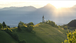 Church with white steeple sits on grassy hillside in alpine valley at sunrise.