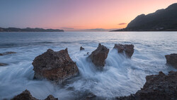 Rocky outcrops surrounded by smooth, flowing seawater at twilight with mountains in the distance.