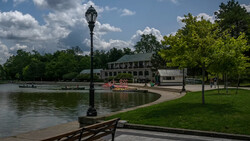 Lamppost on wooden dock overlooking a calm lake with historic building and trees under blue sky.