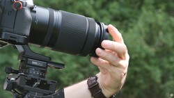 Photographer's hand gripping a telephoto zoom lens mounted on a camera with a tripod head visible below.