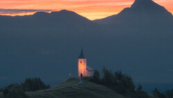 Illuminated church with a steeple on a hilltop at sunset, framed by dark mountains and golden sky.