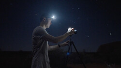 Photographer with headlamp adjusts camera on tripod at night under starry sky.