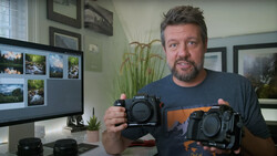 Photographer holding two mirrorless camera bodies side by side in a home office setting.