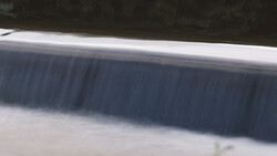 Water flowing over a concrete dam structure with horizontal spillway gates.
