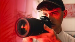 Photographer holding a Nikon camera with a telephoto lens, illuminated by red studio lighting.