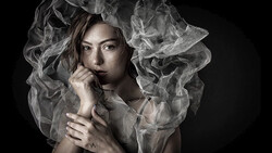 Woman with sheer draped fabric around her head and shoulders against a dark background.