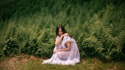 Pregnant woman in white lace dress sitting among tall evergreen trees.