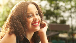 Woman with curly hair smiling outdoors in natural sunlight with blurred foliage background.