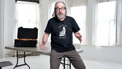 Man in glasses and black t-shirt sitting on table next to open equipment case in bright studio room.