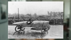 Vintage racing car number 13 on wet track with spectators and trees in background.