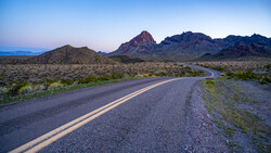 Desert highway with double yellow lines curving toward distant mountains at dusk.