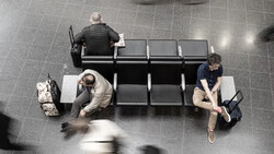 Overhead view of three people sitting on a black bench in a tiled public space.