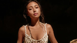 Woman in beaded gold sleeveless top against dark background, looking downward with subtle expression.