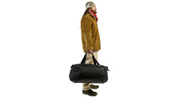 Person in mustard jacket and cap standing against white background while holding black duffel bag.