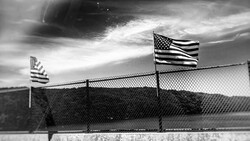 Two American flags on poles behind a chain-link fence under a dramatic cloudy sky, photographed in black and white.