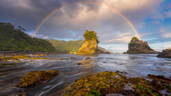 A double rainbow arcs over a dramatic coastal seascape with moss-covered rocks and a prominent sea stack bathed in golden light.