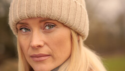 Woman with blonde hair wearing a cream knit beanie, photographed outdoors with soft natural lighting.