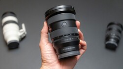 Hand holding a telephoto zoom lens with focus ring and depth-of-field scale visible against gray background.