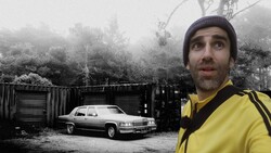 Man in yellow jacket and beanie looking upward beside a vintage car and industrial containers in misty forest setting.