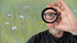 Photographer wearing glasses holds a circular magnifying lens up to his eye, revealing a close-up view of a wildflower meadow behind him.