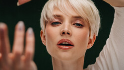 Portrait of a person with platinum blonde pixie cut against a dark background, with hands positioned near their face and shoulder.