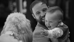 Man holding a smiling toddler next to a fluffy white dog, all three appearing joyful in a black and white portrait.