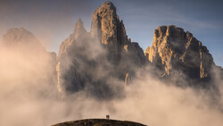 Two hikers standing on a hillside facing towering rock peaks shrouded in mist and clouds.