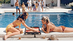 Two women playing backgammon at poolside while guests gather in the background.