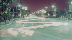 Empty nighttime street intersection with long exposure light trails creating flowing patterns across the pavement.