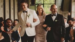 Wedding party walking together down an aisle indoors, with groom in cream blazer, bride in beaded dress, and groomsman in black tuxedo.