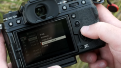 Photographer's hands holding a mirrorless camera with rear LCD screen displaying image quality and file format settings menu.