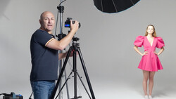 Photographer adjusting camera settings on tripod while model in pink dress poses under studio lighting.