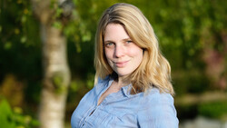 Woman with blonde shoulder-length hair wearing a light blue button-up shirt, photographed outdoors with soft natural lighting.
