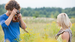Teenage boy photographing a blonde girl in a wildflower field using a vintage film camera.