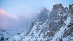 Snow-covered mountain peaks at dawn, with soft pink alpenglow illuminating jagged summits through clearing mist.