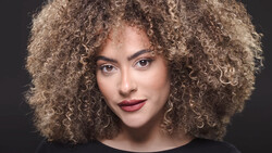 Portrait of a woman with voluminous curly blonde hair against a dark background.