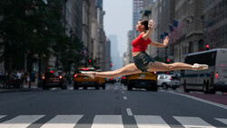Dancer performing a grand jeté across a city street crosswalk with yellow taxis and urban buildings in background.