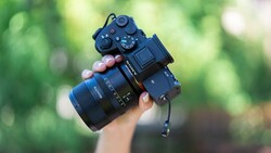 Photographer holding a mirrorless camera with attached lens and external recorder against a blurred green foliage background.