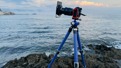 Professional camera on blue tripod mounted on rocky coastline facing calm ocean.