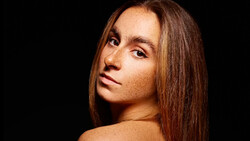 Woman with long brown hair looking over shoulder against black background.