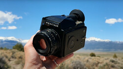 A Pentax 67 medium format camera held in someone's hand against a mountain landscape backdrop.