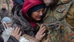 Woman in burgundy winter hat smiling while embraced by two people in military-patterned uniforms.