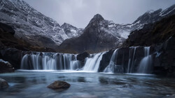 Tiered waterfalls flowing into a blue pool beneath snow-capped mountains at twilight.