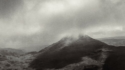 Volcanic cone shrouded in mist under overcast sky with barren lava fields in foreground.
