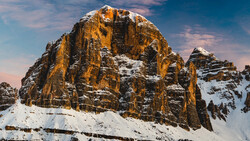 Snow-covered rocky mountain peak with golden-orange columnar formations lit by warm sunlight against blue sky.