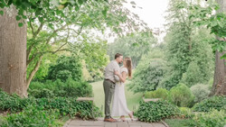 Couple kissing on a stone pathway lined with tall trees and lush green foliage.